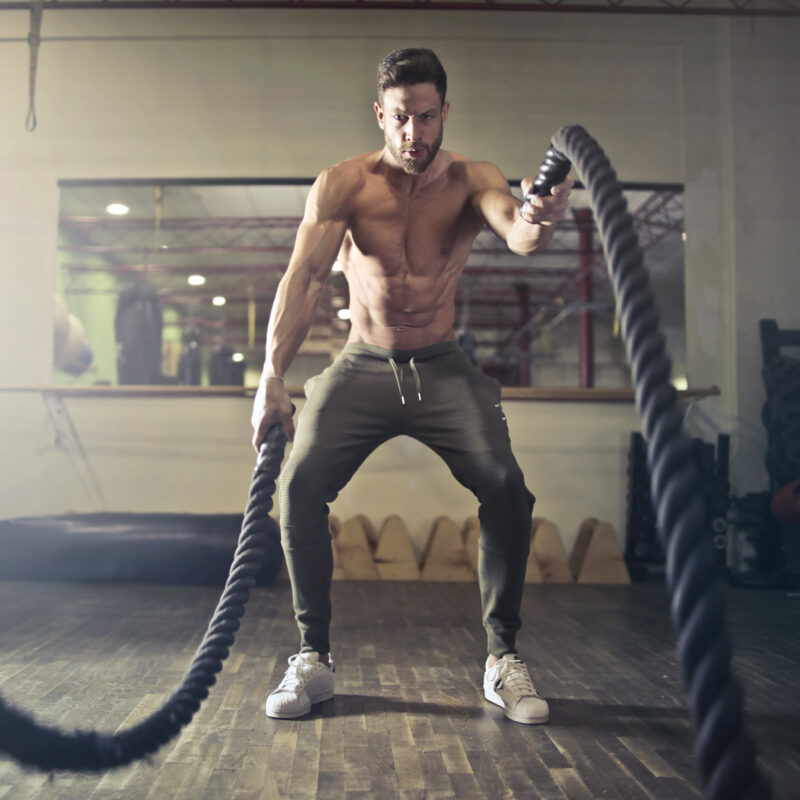 Athletic man exercises shirtless in sweatpants while lifting heavy ropes for weight training.