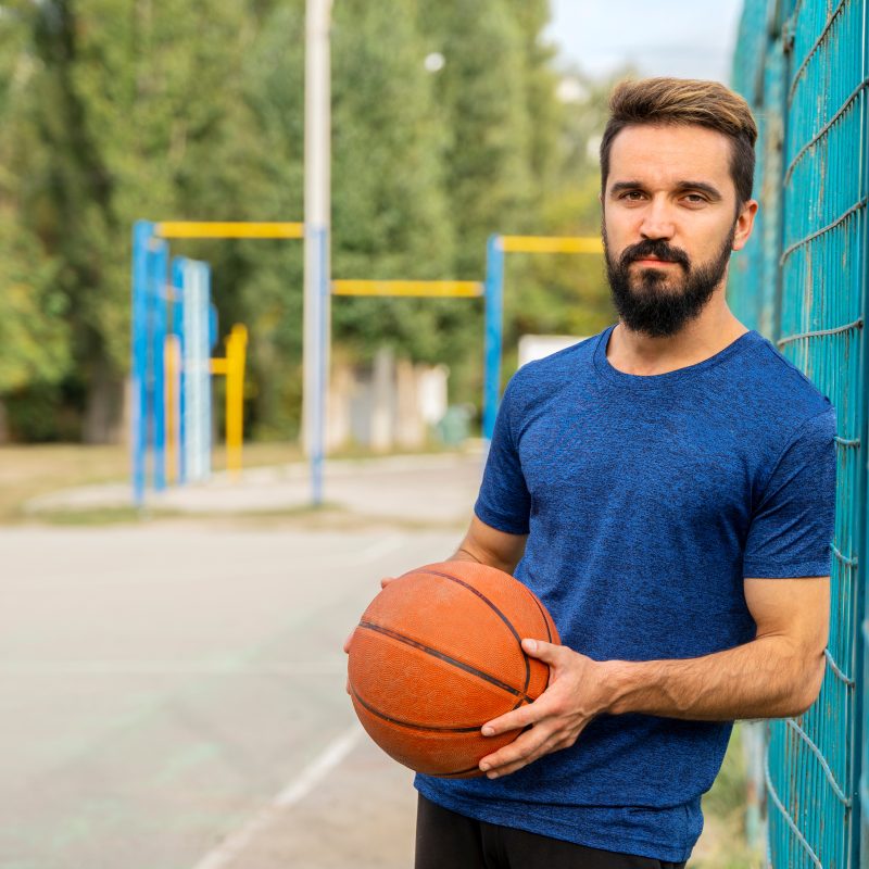 Young brunette man with a basketball ball in his hands and in good health stands against the fence outdoors.